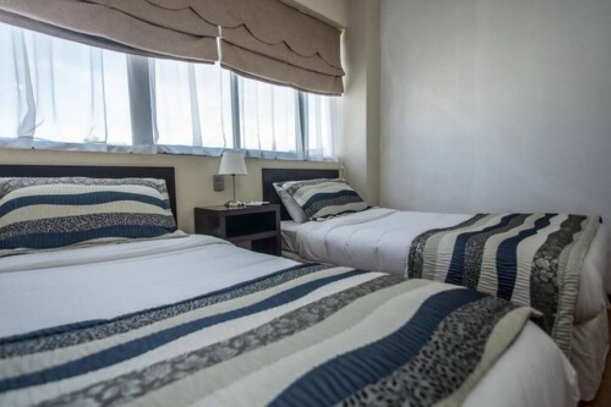 A hotel room with two beds, each with striped bedding, a bedside table with a lamp, and a window with a beige curtain.