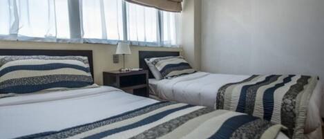 A hotel room with two beds, each with striped bedding, a bedside table with a lamp, and a window with a beige curtain.