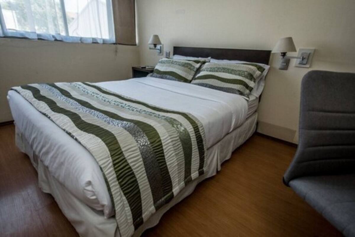 A neatly made bed with striped bedding, a chair, and two wall-mounted lamps in a room with wooden flooring.