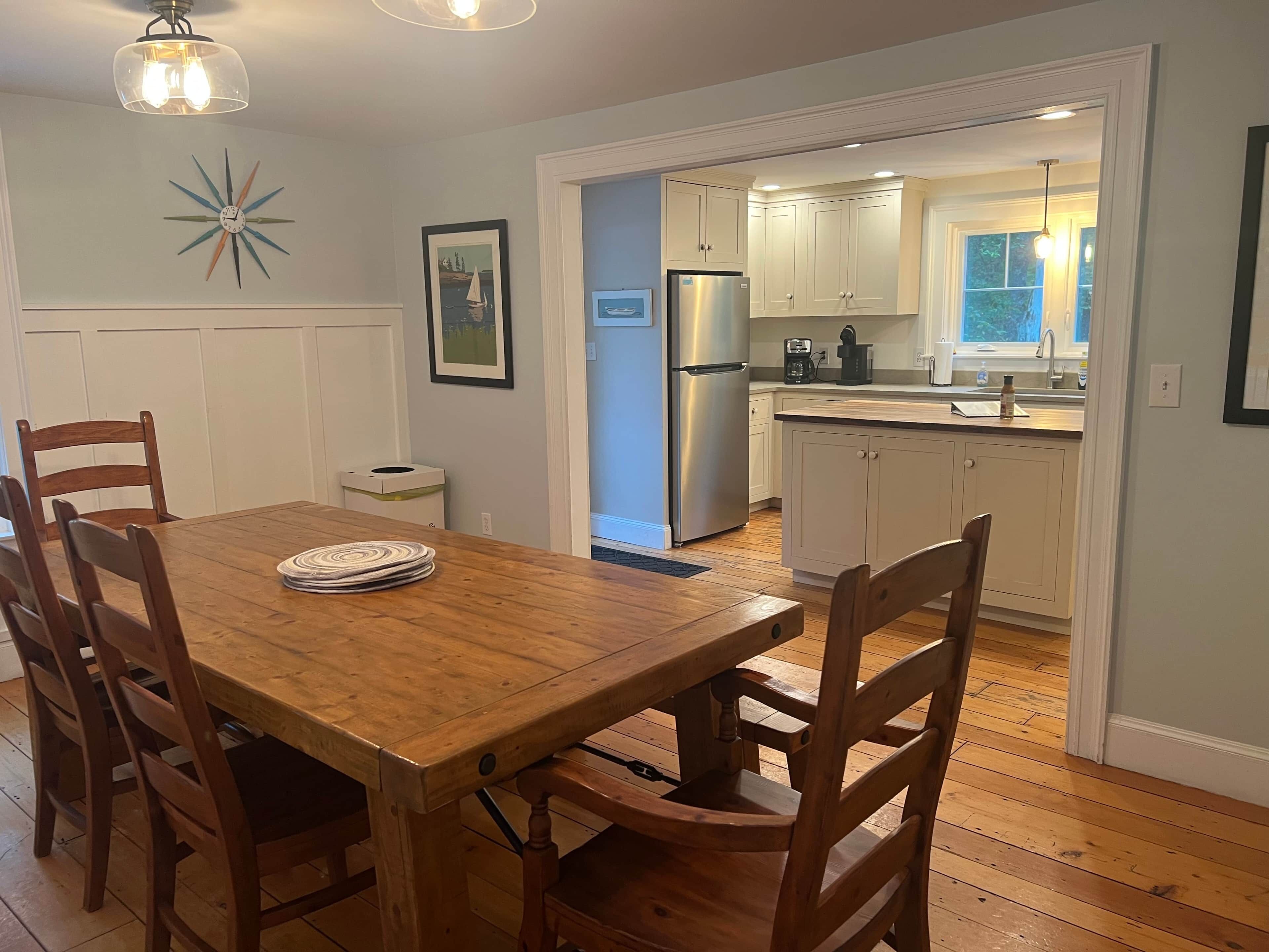 A dining area with a wooden table and chairs, a refrigerator, and a kitchen with white cabinets and a window.