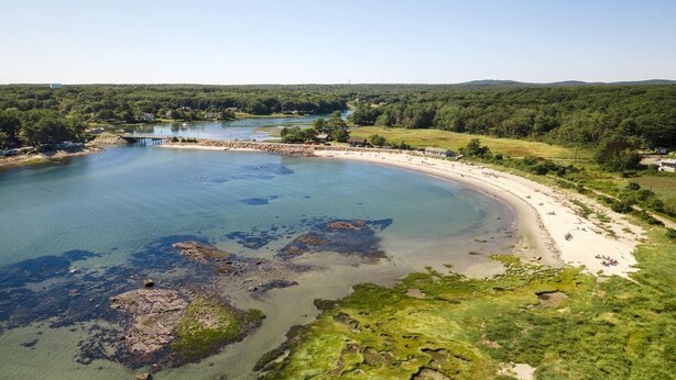 Paesaggio costiero con spiaggia sabbiosa, lago dalle acque limpide e area boschiva sullo sfondo.