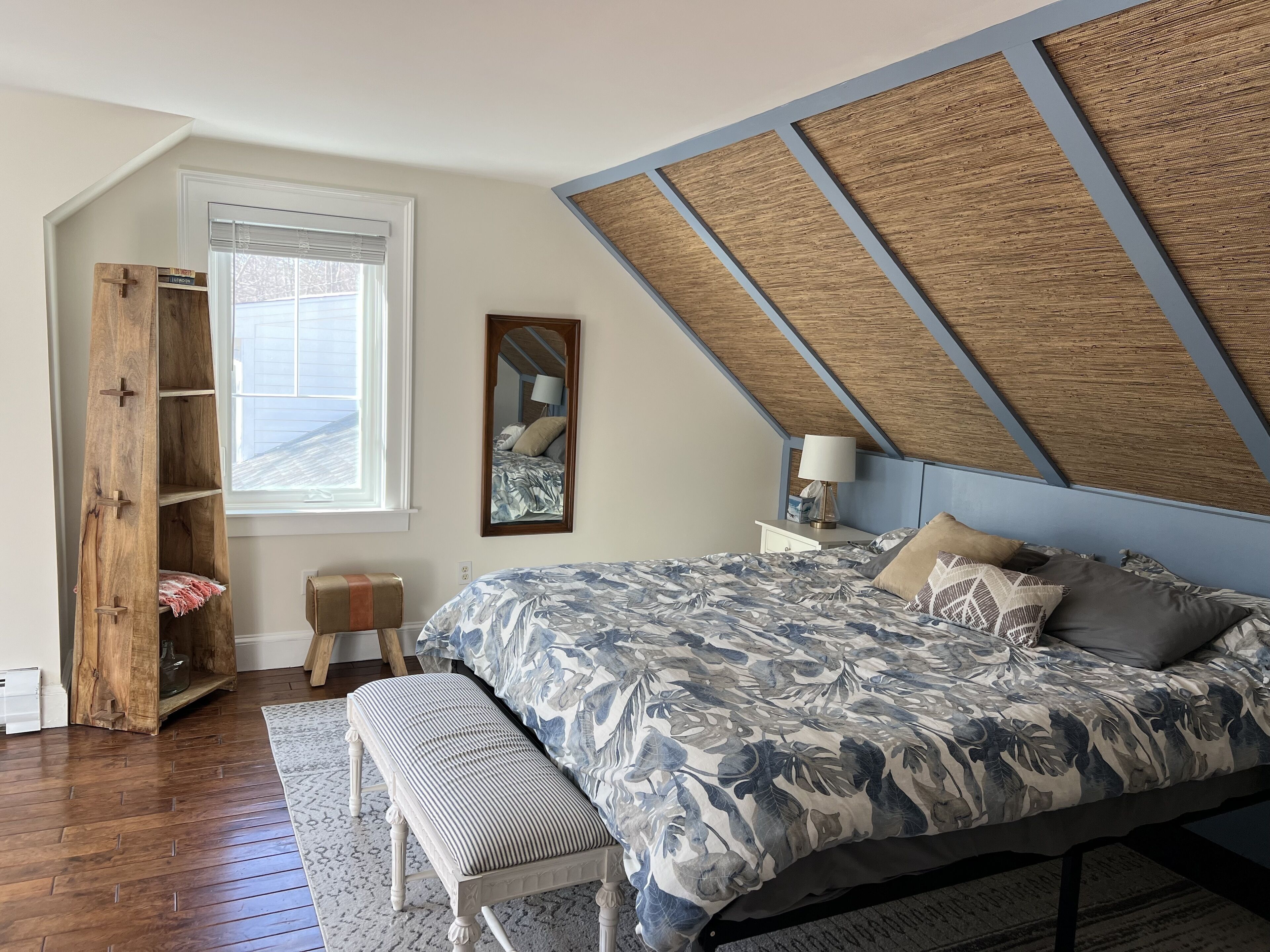 A bedroom with a large bed, a wooden wardrobe, a bench, a nightstand, a mirror, and a window with blinds.