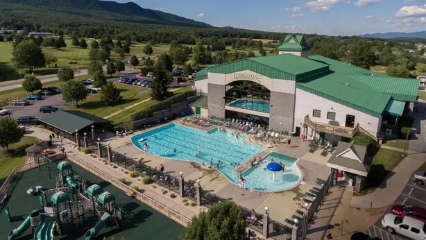 Un hotel con una piscina grande, zona de juegos para niños y montañas al fondo.