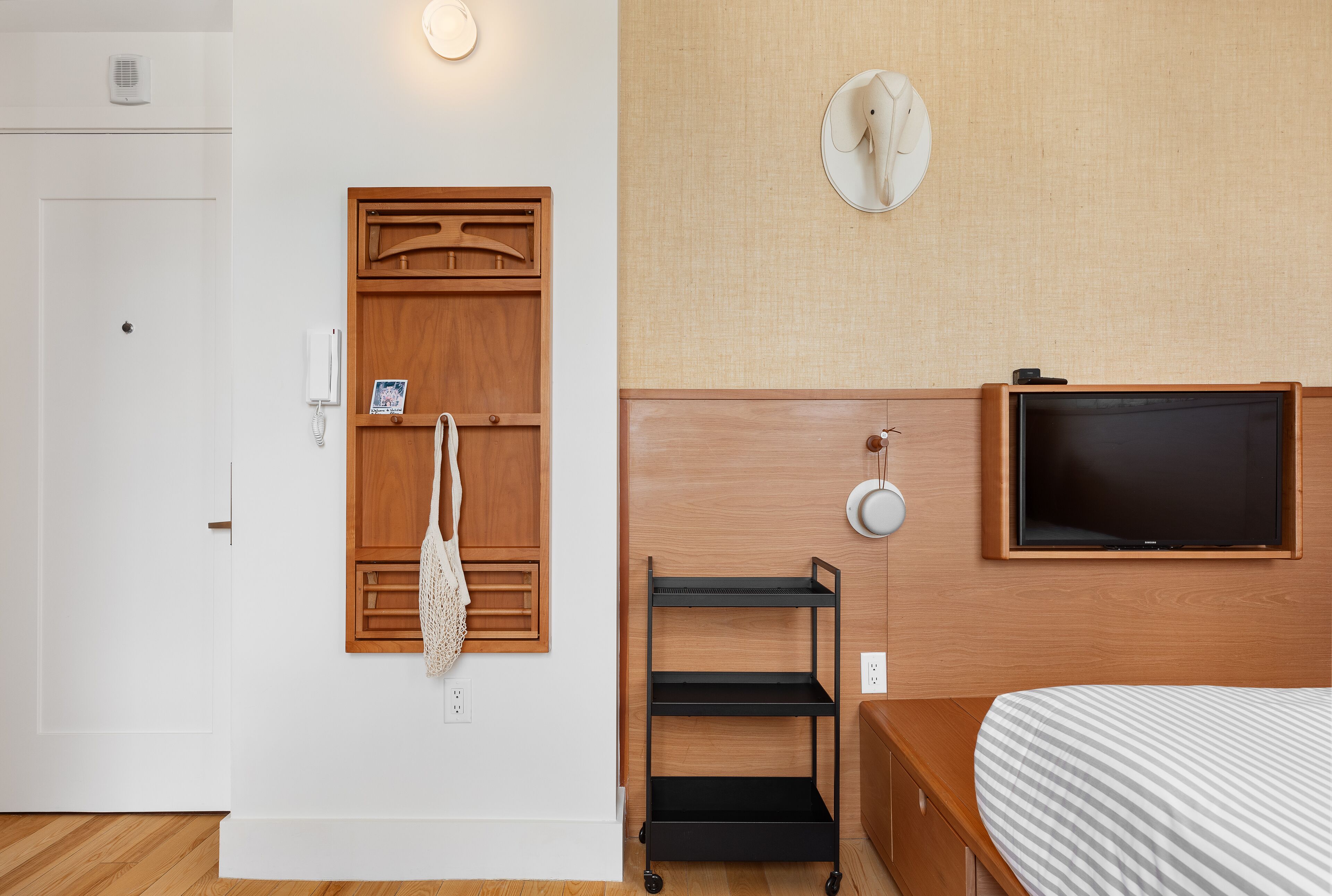 A hotel room with a wooden door, a white wall, a wooden headboard, a TV mounted on the wall, and a small shelf unit.