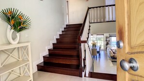 Un escalier en bois menant à un deuxième étage, une console blanche ornée d’une plante et une porte en bois dotée d’une poignée argentée.