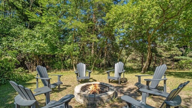 A circular fire pit surrounded by four Adirondack chairs in a forest setting. - Clean and Modern Home near Lk MI (Manistee)