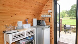 A compact kitchen with a microwave, kettle, and coffee maker. There are mugs on the wall and a small table with glasses and plates. - The Pod at Bank House Farm (Uttoxeter)