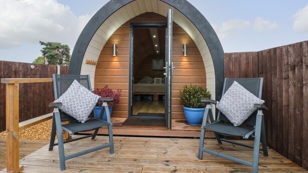 A cozy, semi-open arched structure with a wooden interior, two blue chairs with patterned cushions, and a wooden deck. - The Pod at Bank House Farm (Uttoxeter)