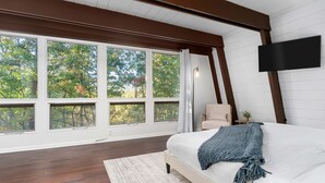 Een slaapkamer met een groot raam, een bed, een stoel en een flatscreen televisie aan de muur.