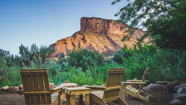 Een houten terras met Adirondack-stoelen en een tafel, omgeven door rotsen en begroeiing, met een berg op de achtergrond.