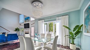 Een moderne woonkamer met een eettafel met glazen blad, witte stoelen, een kroonluchter en een grote potplant.