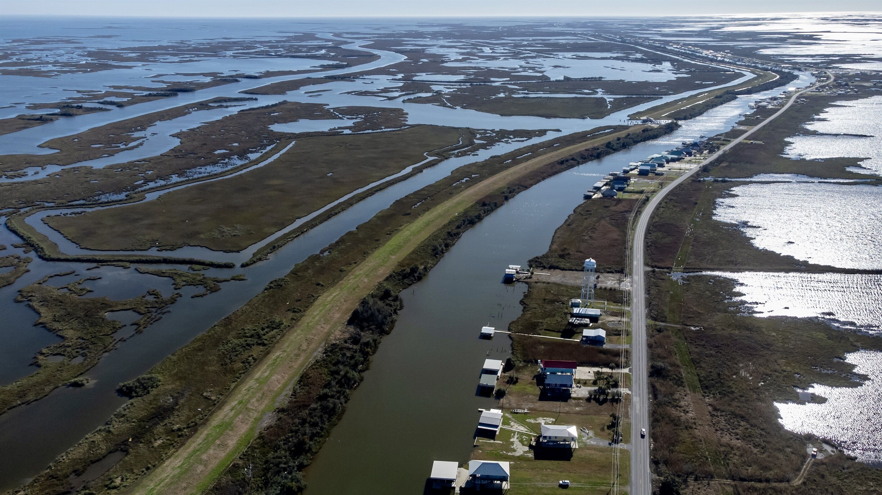 An aerial view of a coastal area with a long, narrow waterway flanked by marshy land and a road with buildings and vehicles along the side.