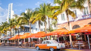 A vintage car parked in front of a restaurant with orange umbrellas and palm trees. - Majestic Mansion • Games, Playground & Pool_BBQ (Miami)