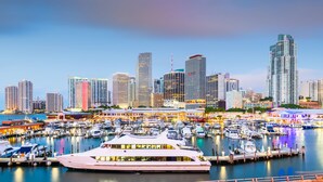 A marina with multiple boats docked, a large yacht, and a city skyline with high-rise buildings in the background. - Majestic Mansion • Games, Playground & Pool_BBQ (Miami)