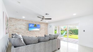 A modern living room with a grey sofa, a ceiling fan, a TV on the wall, and a view of a playground outside. - Majestic Mansion • Games, Playground & Pool_BBQ (Miami)