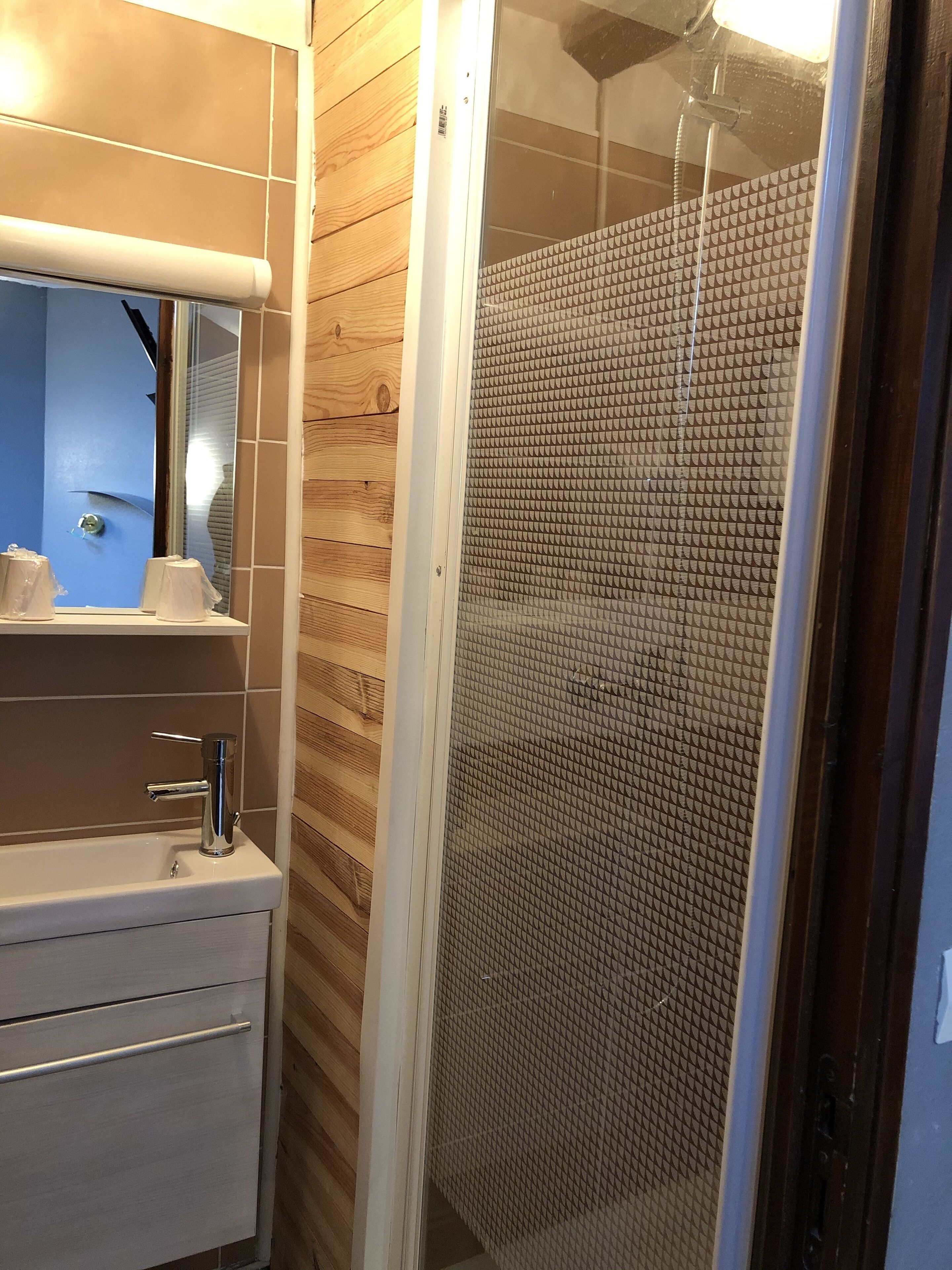 Bany amb paret de fusta, un lavabo amb mirall i una dutxa amb porta de vidre texturat.