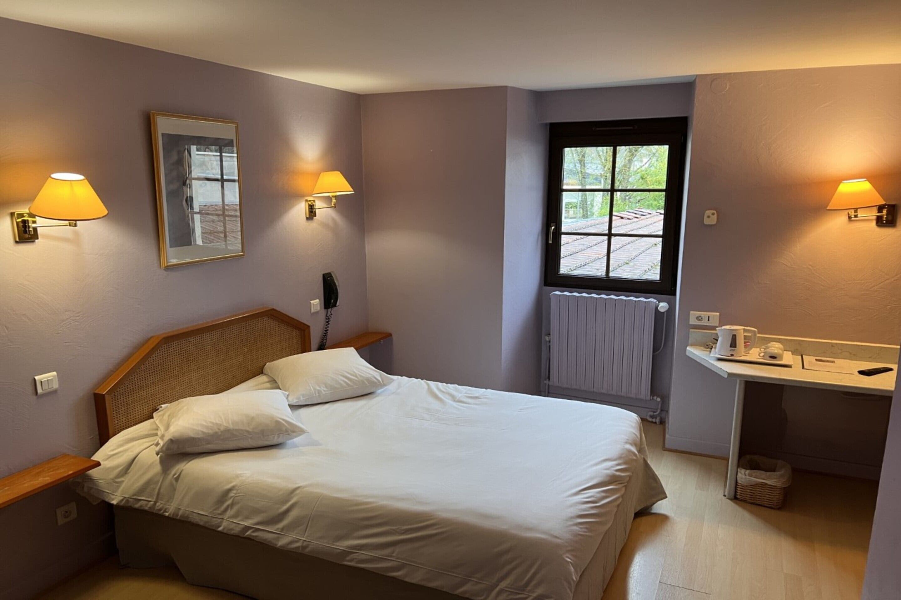 A neatly arranged bedroom with a bed, bedside lamps, a framed picture, a window, and a small table with a kettle.