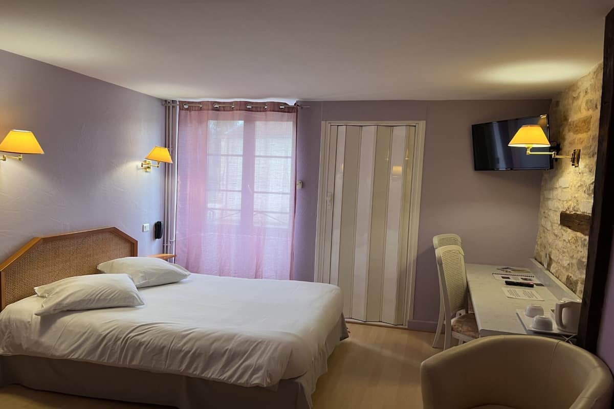Une chambre d’hôtel comprenant un lit, un bureau avec une chaise, un téléviseur fixé à un mur en pierre et une fenêtre avec des rideaux.