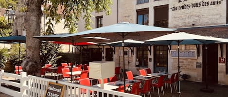 An outdoor café with red chairs and umbrellas, a signboard indicating it's open on specific days, and a building with a sign in a foreign language.
