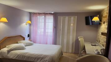 A hotel room with a bed, a desk, a chair, a TV mounted on a stone wall, and a window with curtains.
