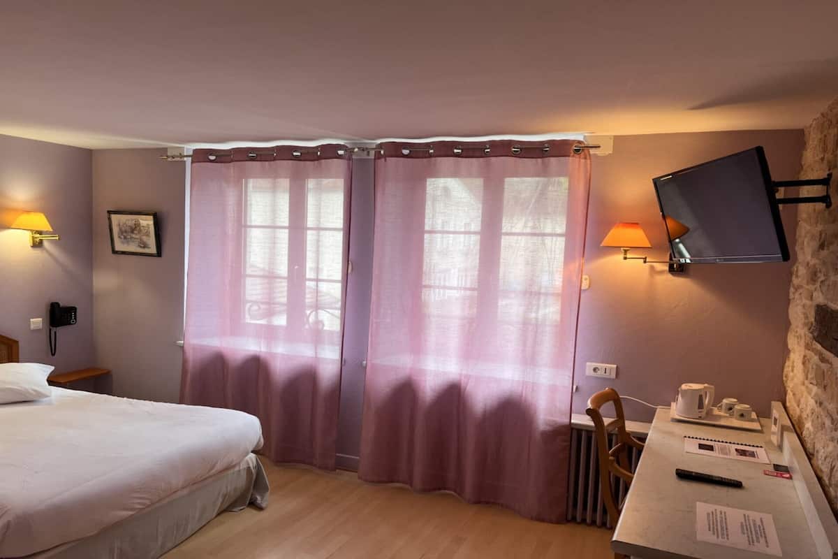 Une chambre d’hôtel avec un lit, un bureau avec une lampe, une télévision et une fenêtre avec des rideaux.