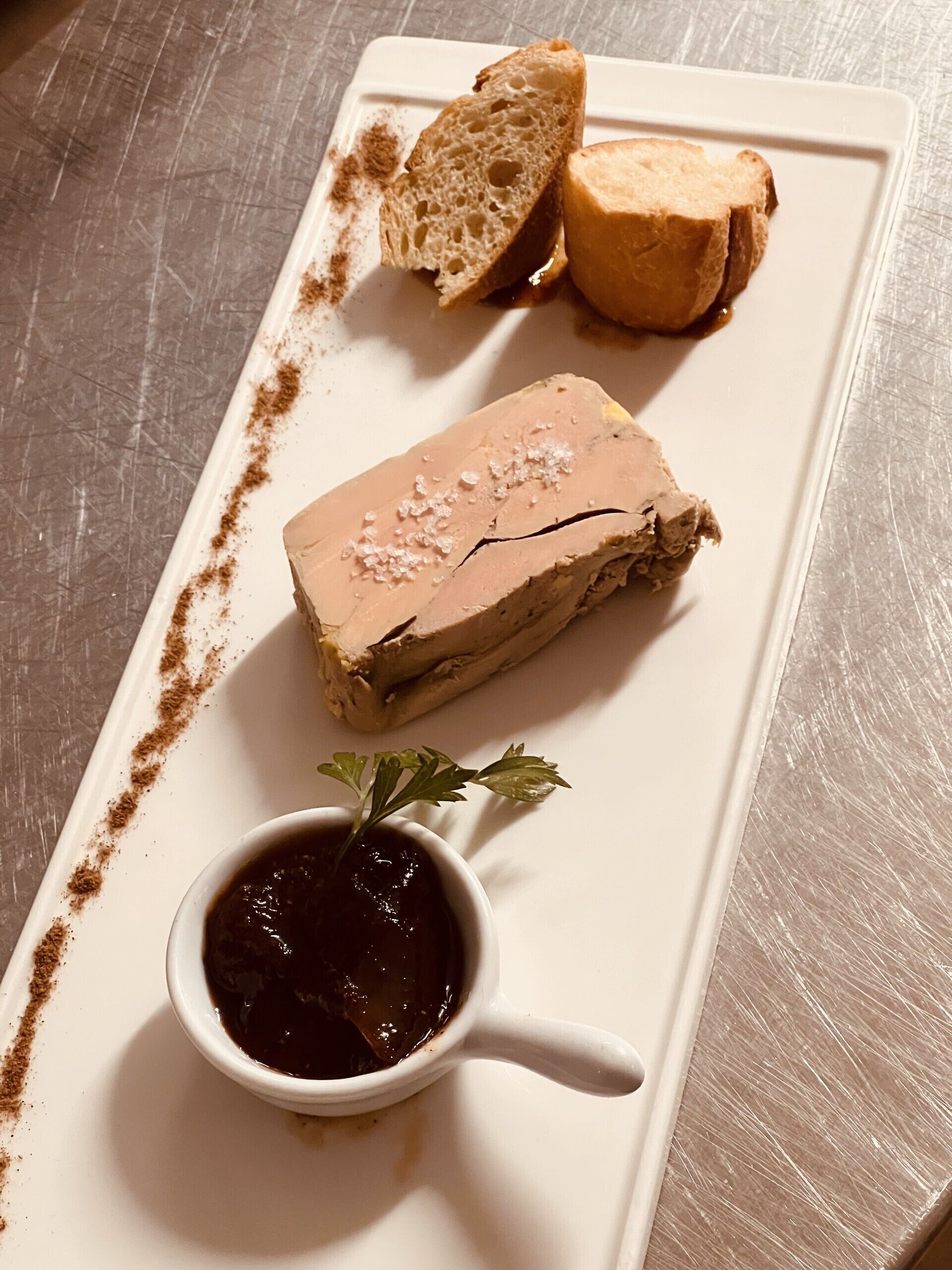Un plateau de service rectangulaire blanc, sur lequel on trouve une portion de terrine, deux tranches de pain et un petit bol de sauce, agrémenté d'une branche de persil.