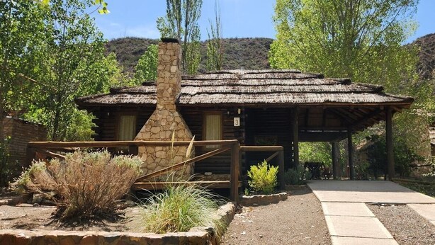 A rustic cabin with a stone chimney, surrounded by desert vegetation and a mountain backdrop.