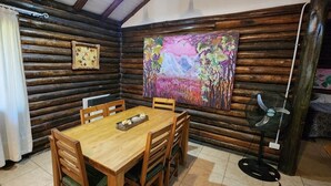 A wooden cabin interior with a dining table, chairs, a painting, and a fan.