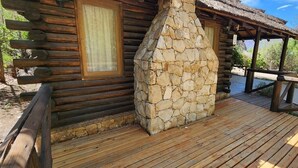 A log cabin with a stone chimney, wooden deck, and a thatched roof structure.