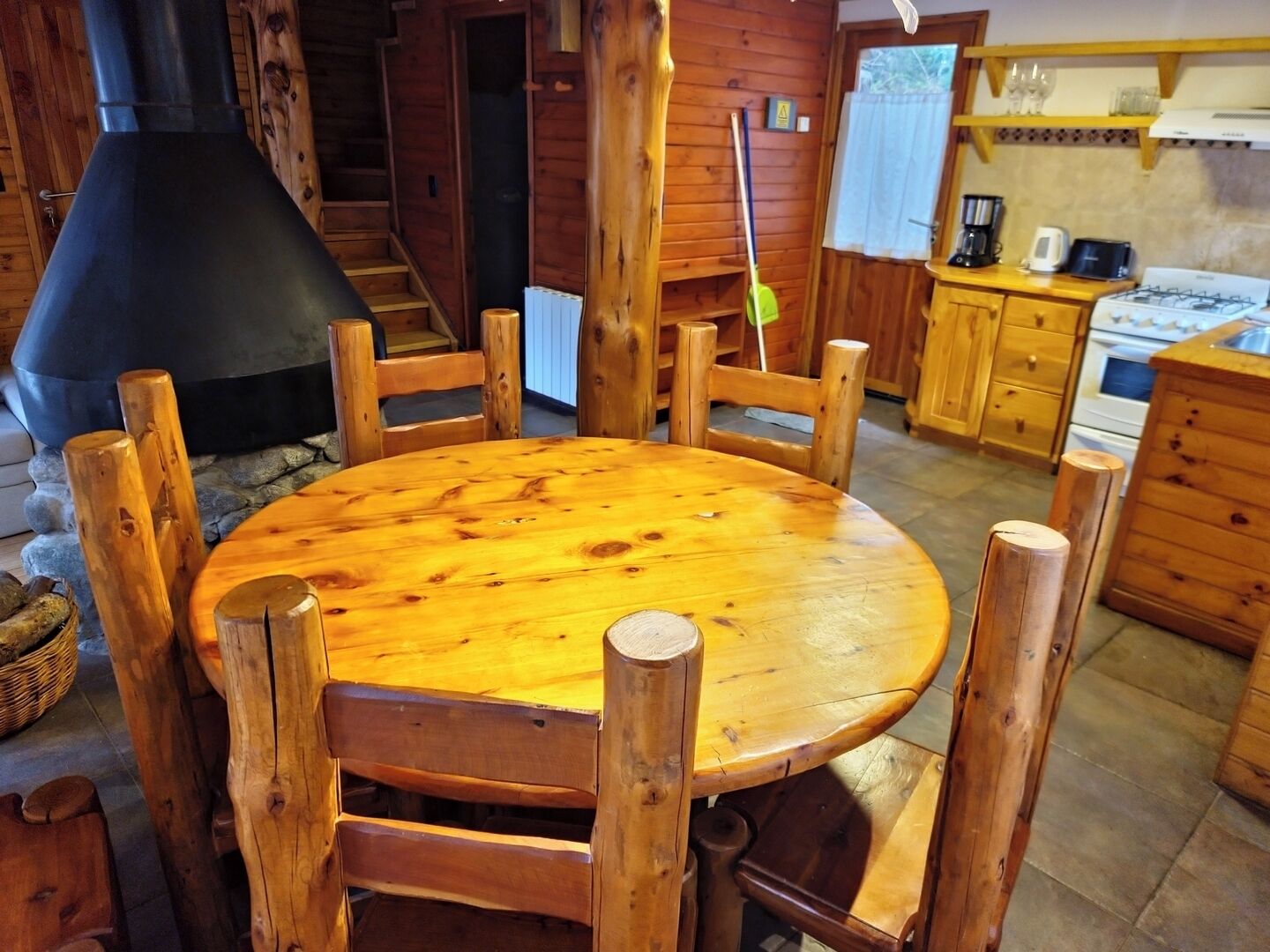 A wooden dining table with chairs, a kitchen area with wooden cabinets, a stove, and a chimney.