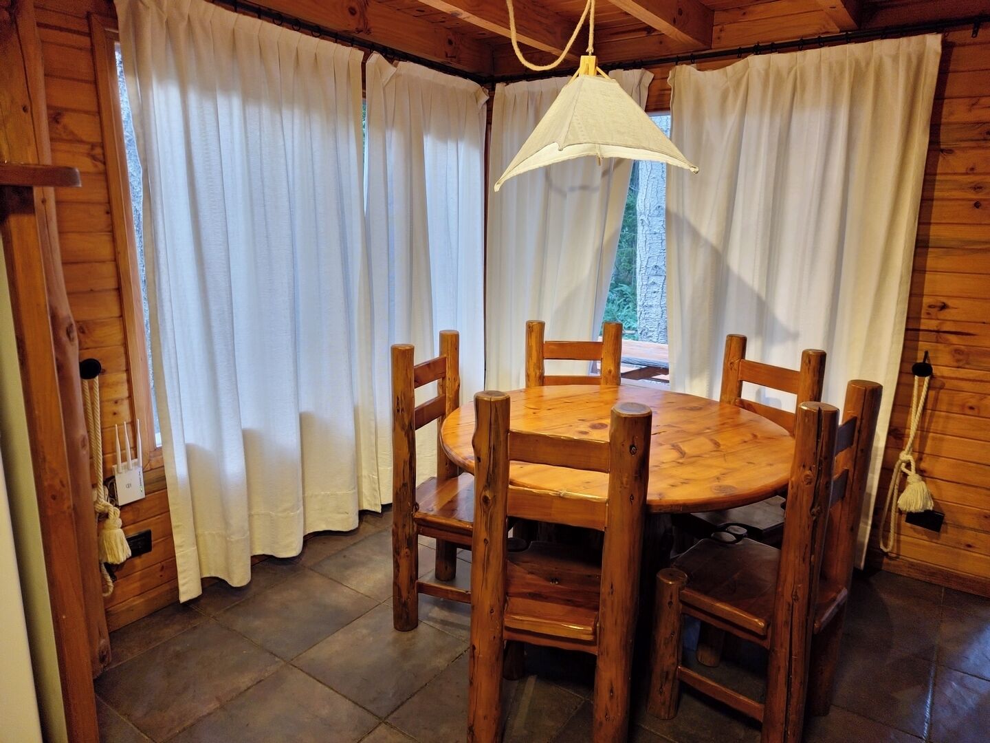 Comedor de madera con una mesa redonda y seis sillas, cortinas blancas y una lámpara colgante.
