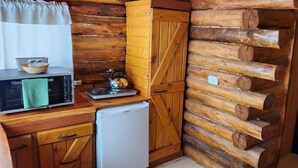 A compact kitchen with wooden log walls, a microwave, a small refrigerator, and a kettle on the counter.