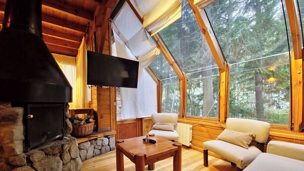 A cozy cabin interior with a stone fireplace, wooden furniture, and large windows offering a forest view. - El Remanso - Radal Cabin, Manzano Bay (Puerto Manzano)