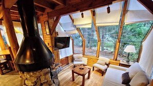 A cozy cabin interior with a stone fireplace, wooden furniture, and large windows offering a view of the forest. - El Remanso - Radal Cabin, Manzano Bay (Puerto Manzano)