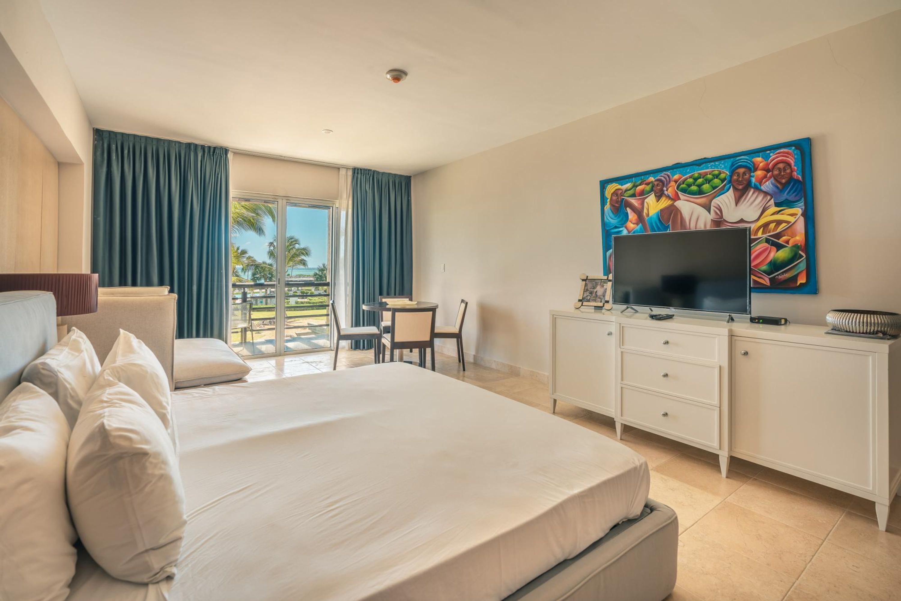 A hotel room with a large bed, a desk with a TV, a painting on the wall, and a balcony with a view of palm trees.