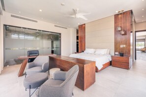 A modern bedroom with a large bed, a desk, and a chair. There is a glass wall and a ceiling fan.