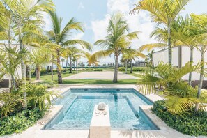 A swimming pool with a stone sculpture, surrounded by palm trees and a view of the ocean.