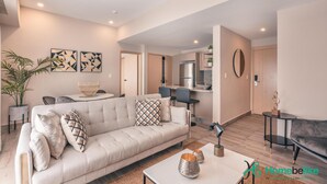 Ein modernes Wohnzimmer mit einer Couch, Couchtisch und einem Essbereich mit Stühlen. Im Hintergrund ist eine Küche mit Kühlschrank und Schränken zu sehen.