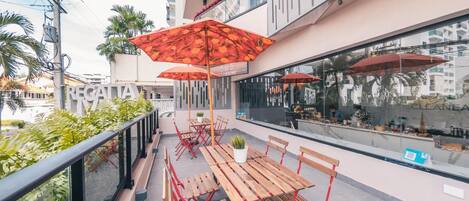 A balcony with wooden tables and red umbrellas, overlooking a cityscape.