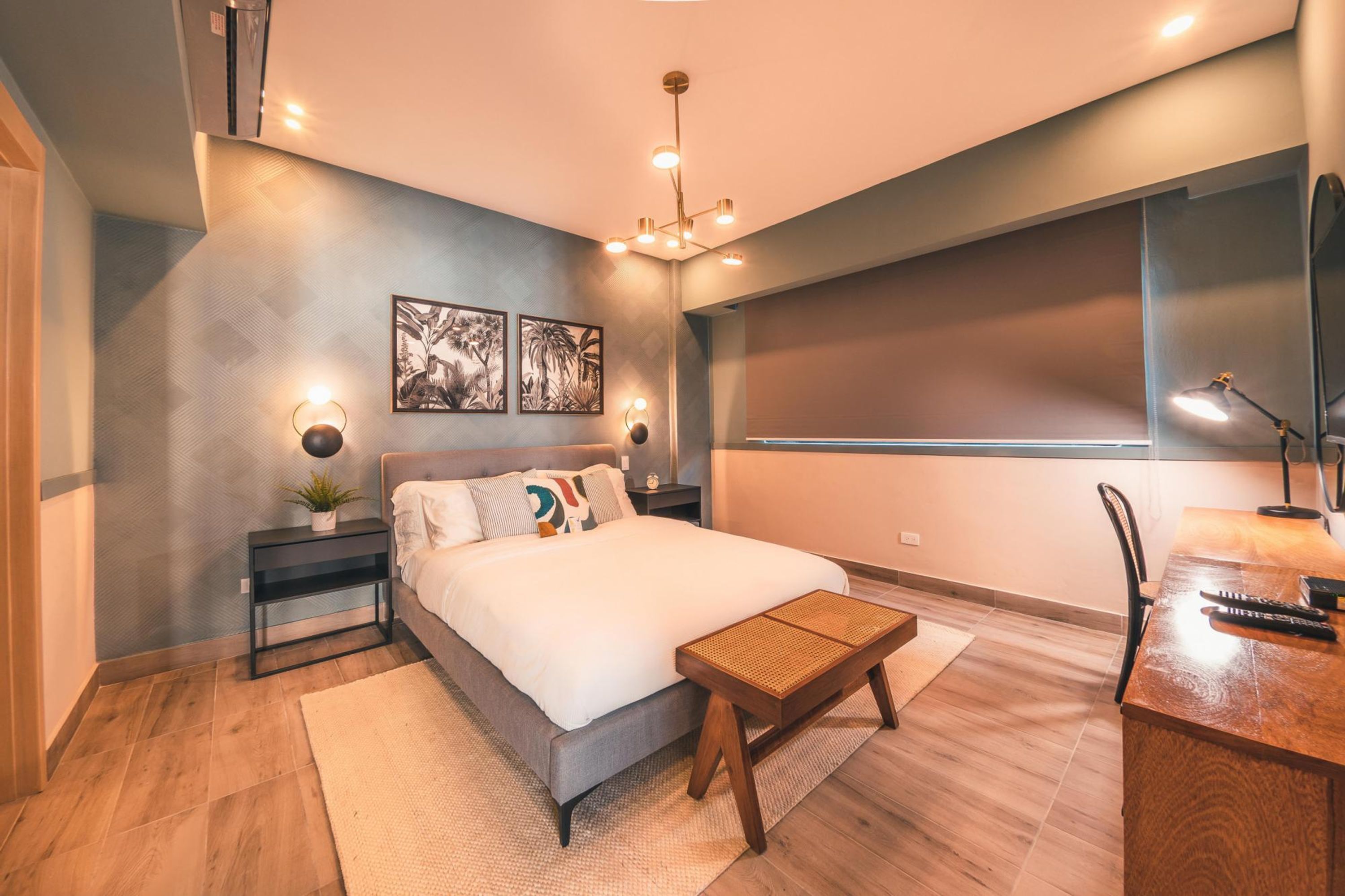 A modern bedroom with a large bed, bedside tables, a desk, and a chair. There are wall-mounted lights and a ceiling light fixture.
