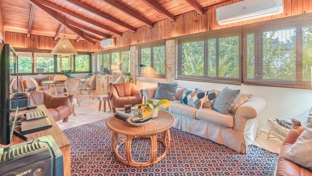 A spacious living area with a high ceiling, wooden beams, a large window with a view of trees, a sofa with cushions, a coffee table, and a dining area with chairs and a table. - 5BR 14 Guests Maid Included Casa de Campo (La Romana)