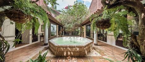 A serene indoor garden with a central hot tub, surrounded by lush greenery and traditional architecture.