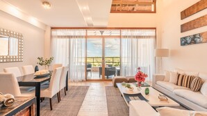 Amplia sala de estar con comedor, sofá y mesa de centro. Cuenta con un ventanal con cortinas, un candelabro y vistas al exterior.