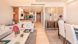Una sala de estar moderna con un sofá blanco, una mesa baja con flores y un comedor con sillas blancas y un aparador de madera.
