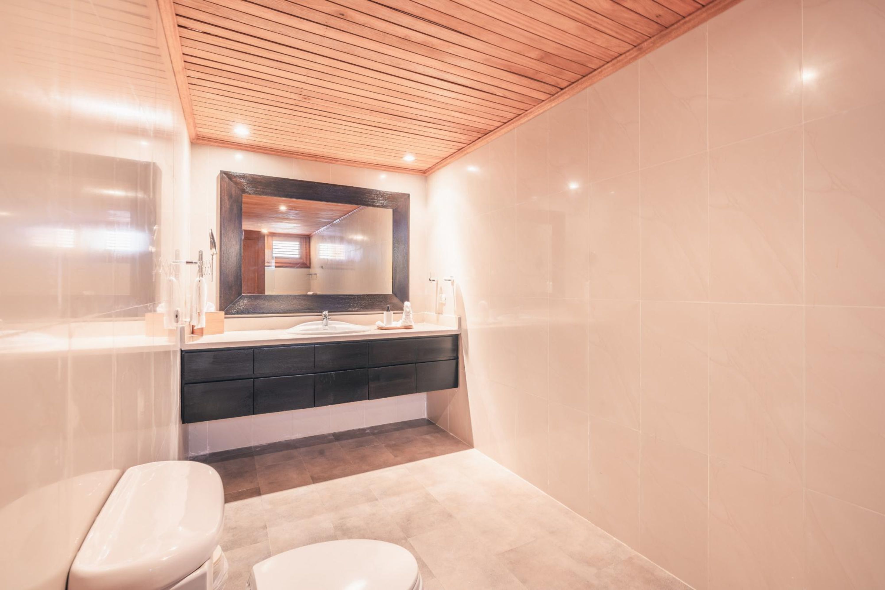 A modern bathroom with a wooden ceiling, a large mirror, a dark vanity, and a toilet.