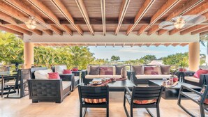 An outdoor patio with wooden ceiling beams, cushioned seating, and a view of palm trees. - Wonderful Villa w Style at Colinas Casa de Campo (La Romana)