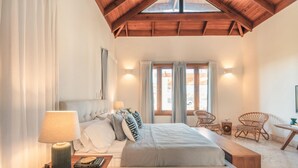 Een slaapkamer met een bed, nachtlampjes, een leestoel, een klein tafeltje en een raam met gordijnen.