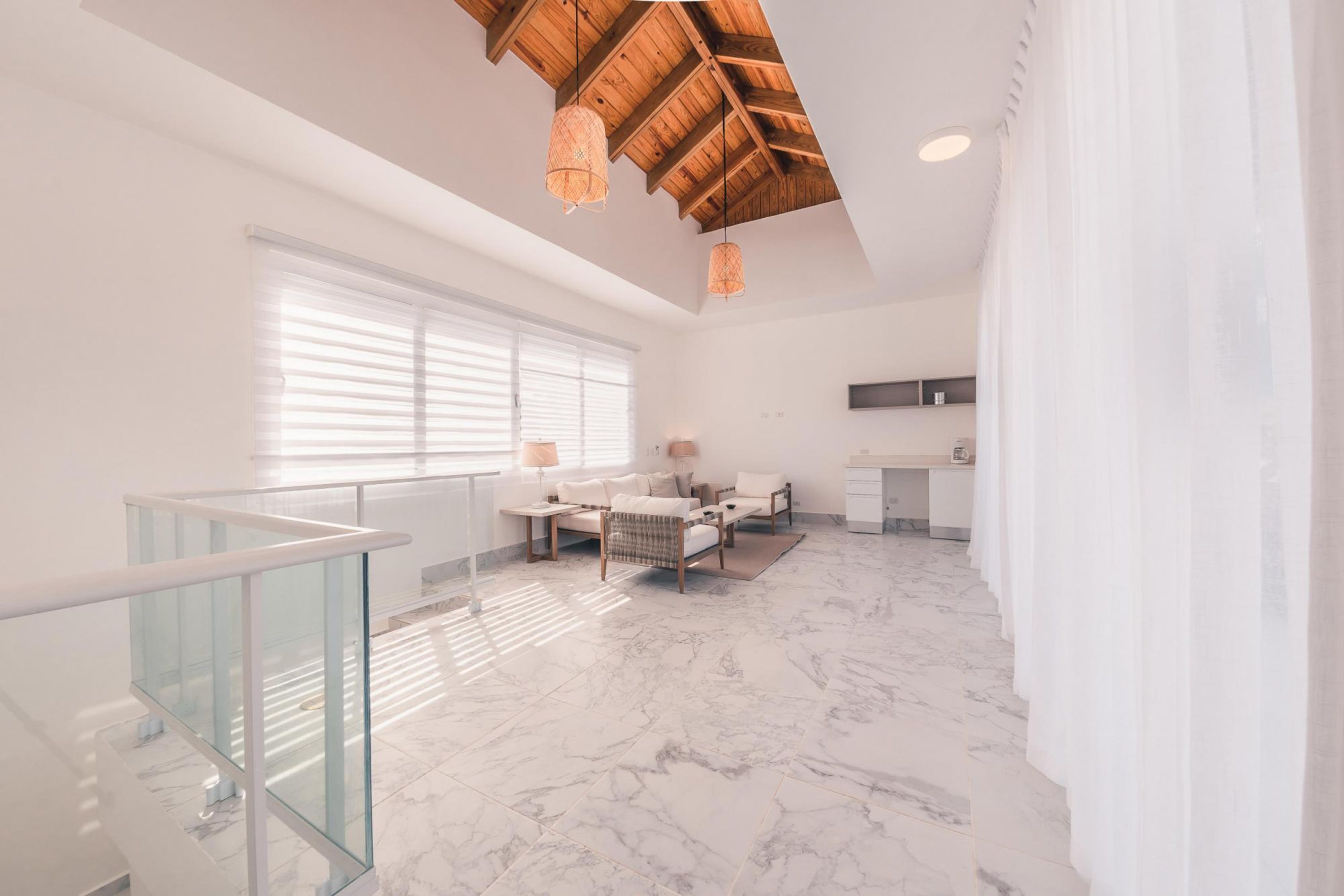 A spacious living area with a high ceiling, a glass railing, a sofa set, a dining table, and a kitchen area.