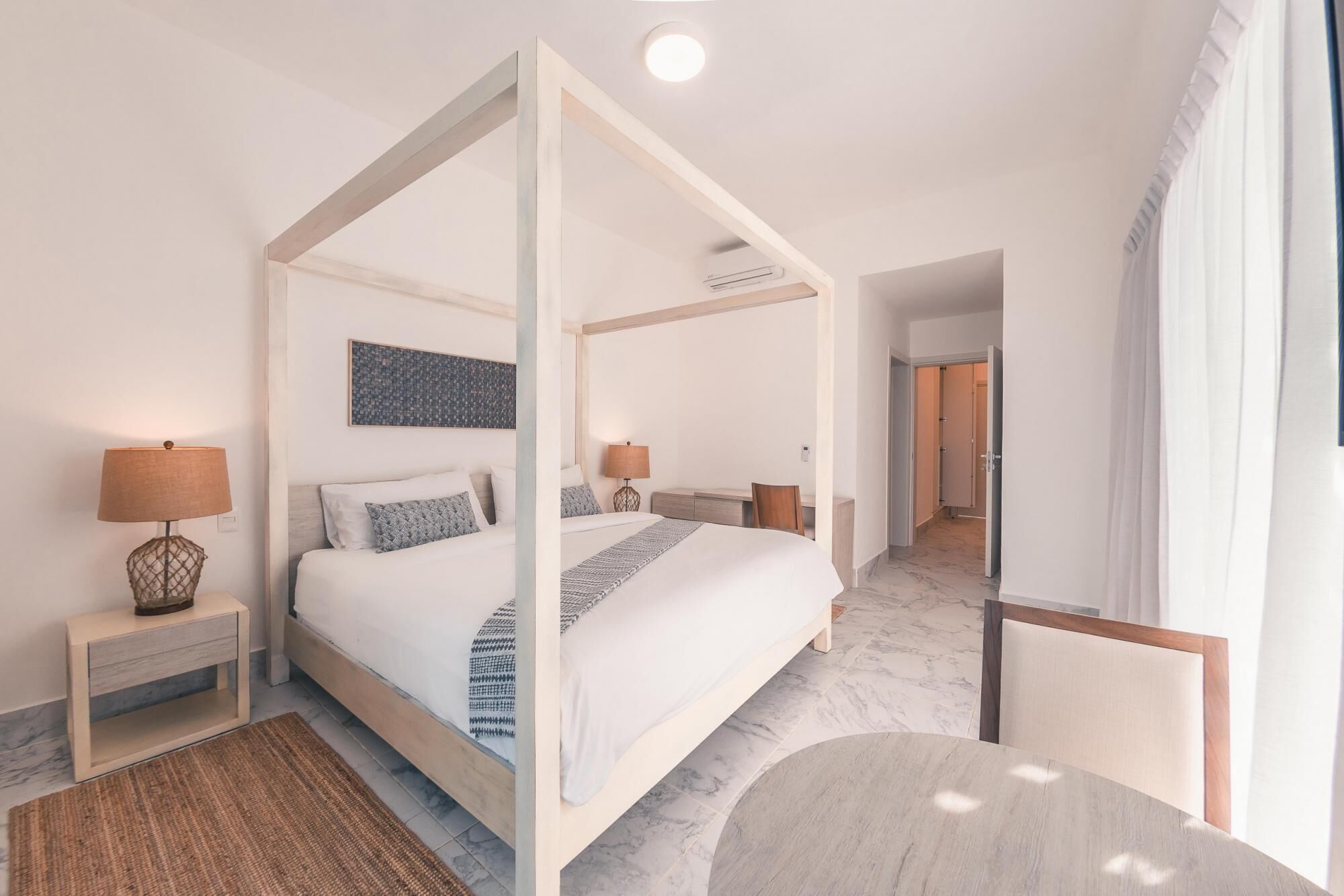 Una cama moderna con dosel sobre un suelo de mármol, mesitas de noche con lámparas y vistas a un pasillo.