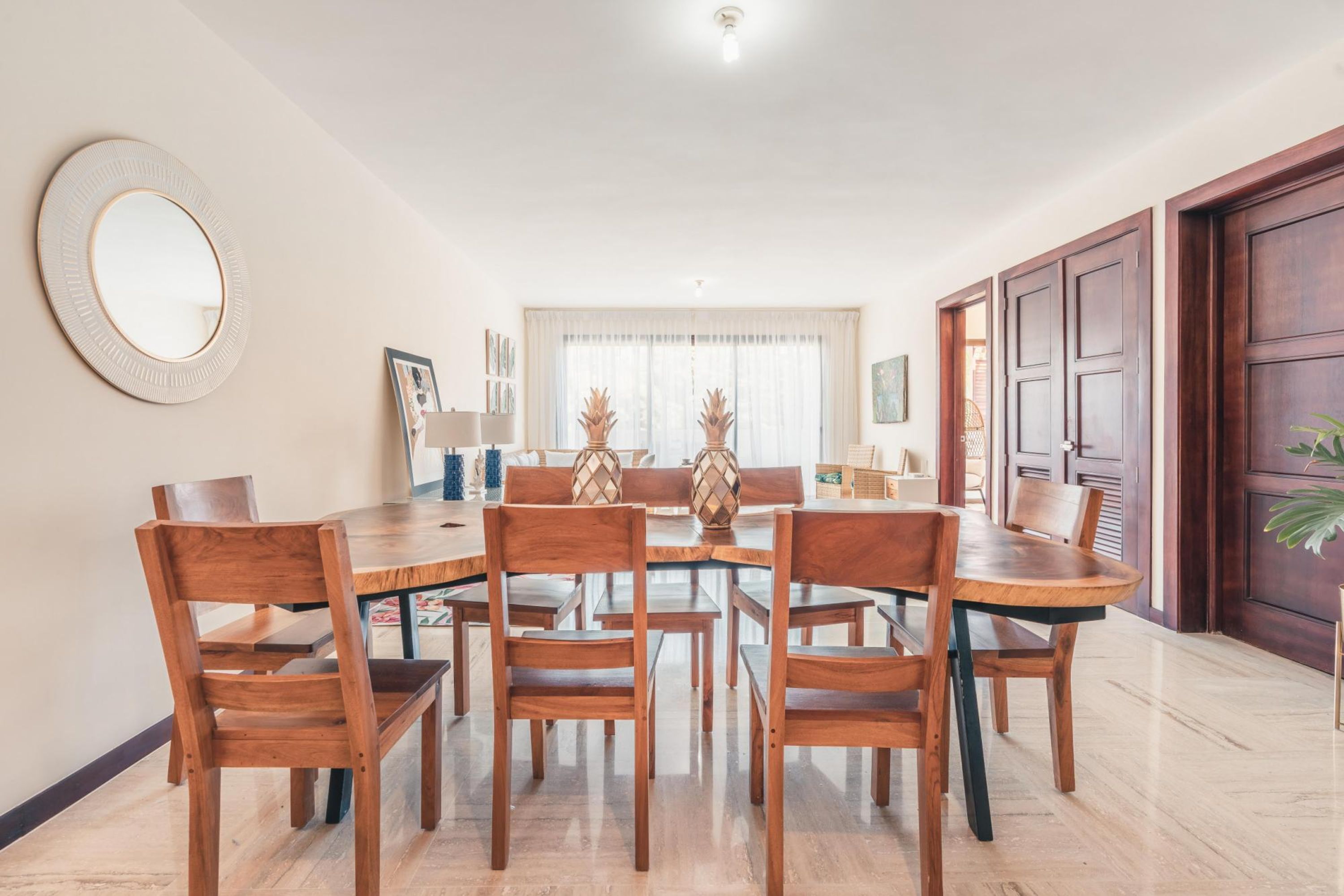 A dining area with a round table and wooden chairs, a large mirror on the wall, and a window with curtains.